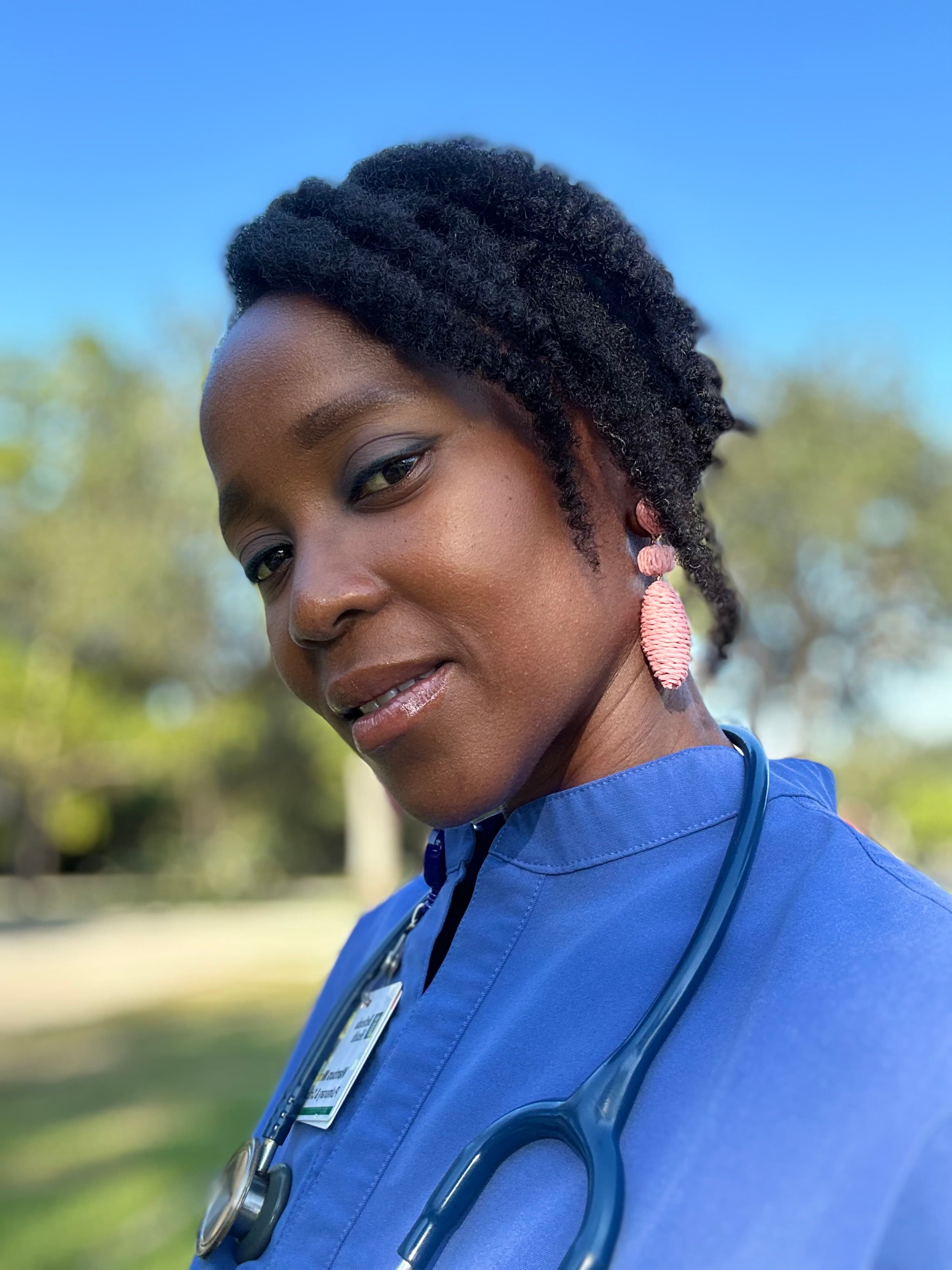 Smiling Black female healthcare professional in blue scrubs with a stethoscope, standing outdoors.